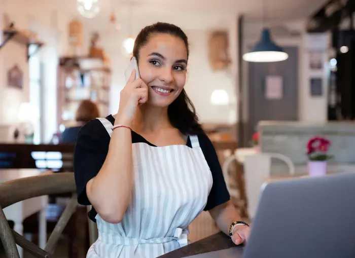Cheerful cafe owner in apron talking on phone while working on laptop inside cozy modern cafe